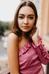 Pensive girl calmly looks into camera, posing for street photo. Cute model in stylish top with frills straightens her hairdo