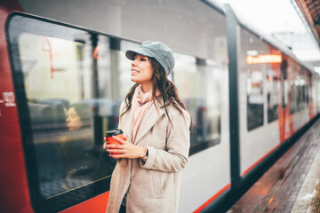 Pretty woman holding red cup and enjoying coffee during waiting the train at the station. Girl Enjoying winter morning under the snowfall with hot drinks and listening to music on headphones.