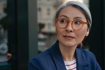 Close up portrait of pensive mature businesswoman looking away, planning start up. Beautiful middle aged woman wearing stylish eyeglasses standing outdoors, focus on face