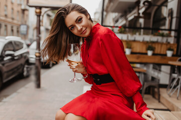 Contented, sweet girl smiles gently and looking into camera. Red dress adds brightness for outfit of lady posing with glass of wine