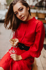 Deep gaze of young woman�s with brown eyes attracts attention. Lady in silk red dress holding glass of french wine