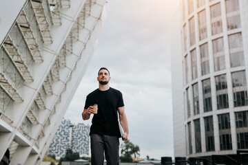 young handsome brunette man walking down the street, carrying his laptop while drinking coffee on the go, lunch break, busy days, multitasking