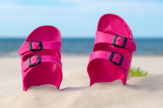 Pink sandals at the beach on a beautiful sunny day. Slippers in the sand by the sea. Flip flops at the shore by the ocean.