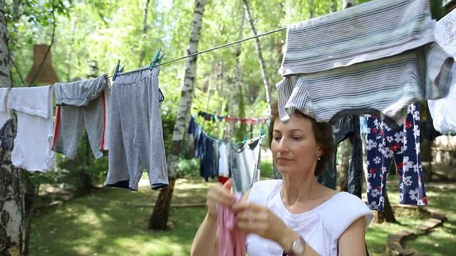 Candid real life portrait of young adult beautiful attractive caucasian woman hanging up fresh washed family clothes on birch tree clothesline with pins at home yard on bright sunny day outdoors