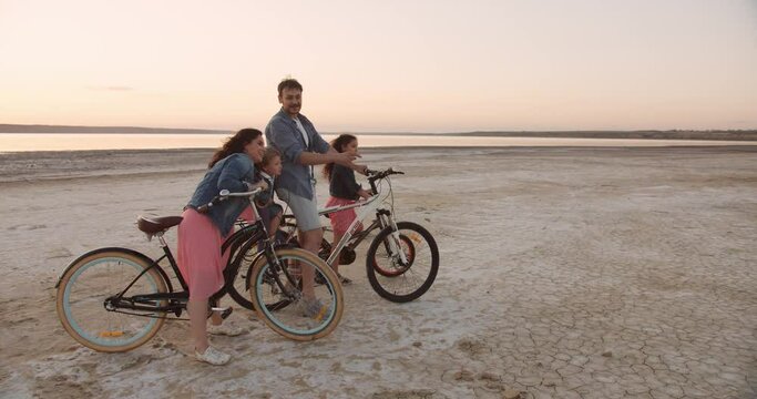 Family of four - mother, father, daughter and little son riding bicycles near the lake during beautiful sunset. Summer vacations concept