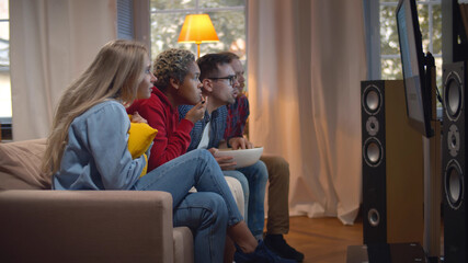 Young diverse people relaxing on couch at home while watching movie and eating popcorn