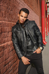 A young handsome stylish guy in a black leather jacket is smiling and leaning against the red brick wall outside.