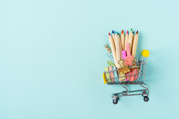 Buying school supplies, pencils, tape, notebook and crayons. Stationery for kids in a shopping cart on blue turquoise background, copy space. Back to school concept. Flat lay top view