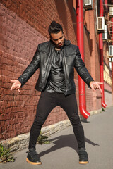 Young handsome stylish guy dancer in a black leather jacket on the street near a red brick wall.