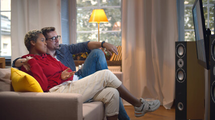Side view of happy young diverse couple indoors at home sitting on sofa hugging and watching TV