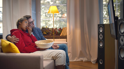 Side view of multiethnic young couple relaxing on couch with bowl of popcorn and watching tv