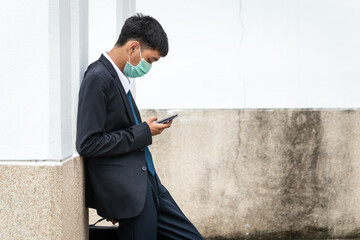 A handsome young asian business man using smartphone. Asian young attractive businessman wearing a face mask leaning against the wall.