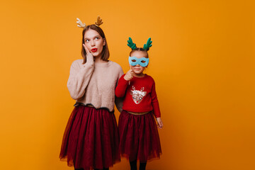 young girl stares in surprise, holding her cheek up while her little sister looks intocamera through the holes in carnival mask.