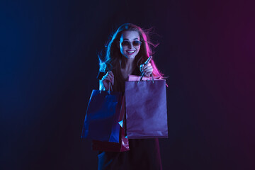 Astonished, shocked with shopping bags, cheerful. Portrait of young woman in neon light on dark backgound. The human emotions, black friday, cyber monday, purchases, sales, finance concept.
