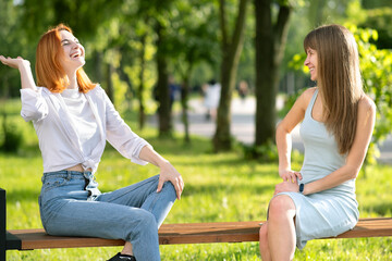 Two young girls friends sitting on a bench in summer park chatting happily having fun.