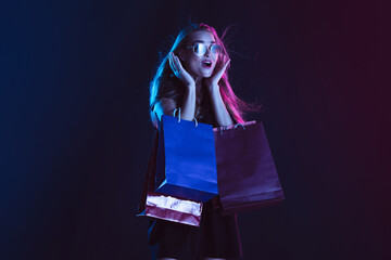 Astonished, shocked with shopping bags, cheerful. Portrait of young woman in neon light on dark...