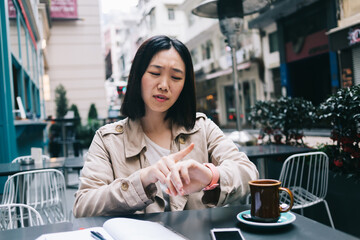 Upset modern Asian woman looking at wristwatch while sitting at street cafe