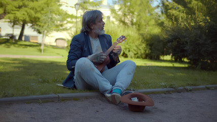 Street musician beggar playing guitar asking for money or food sitting on grass in park