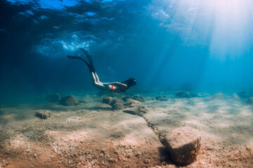 Slim woman in bikini glides at blue sea with sun rays. Freediving with fins underwater in ocean