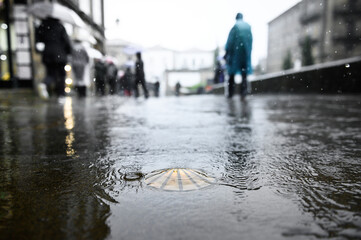 Bronze scallop as a waymark in the old town of Santiago de Compostela on wet ground during rain with pilgrims in ponchos in the background.