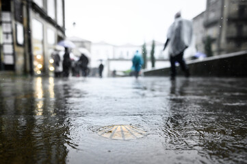 Bronze scallop as a waymark in the old town of Santiago de Compostela on wet ground during rain with pilgrims in ponchos in the background.