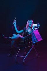 Rolling in a cart with shopping bags and megaphone. Portrait of young woman in neon light on dark...