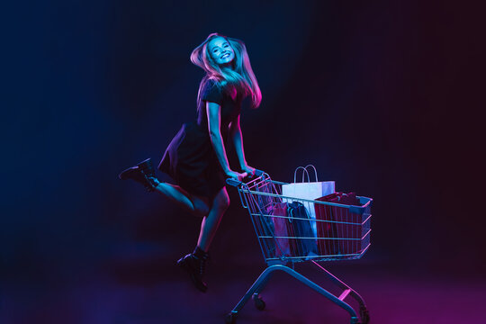 Rolling in a cart with shopping bags. Portrait of young woman in neon light on dark backgound. The human emotions, black friday, cyber monday, purchases, sales, finance concept. Trendy colors.