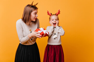 The little girl is glad and surprised by the gift in the red box with a white bow. A young mother gives her daughter a Christmas gift and looks at her reaction.