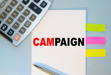 CAMPAIGN is written on a notepad next to multicolored stickers, a calculator and a pen on a blue background. Business concept