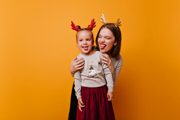 Cool Mom and her active daughter, in ears of New Year's deer of different colors, having fun in front of camera showing their tongues