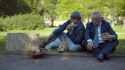 Successful businessman sitting on ground with homeless beggar and eating pastry together in park