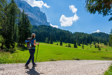 Young woman with a hat, trekking poles, a backpack hiking on a trail through green meadows in the...