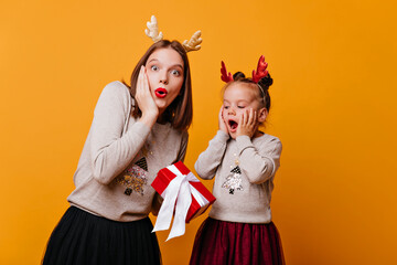 Mom gives her little daughter a gift in a red box on an isolated orange background. A young woman with her daughter in identical Christmas sweaters is looking at the camera in surprise and delight.