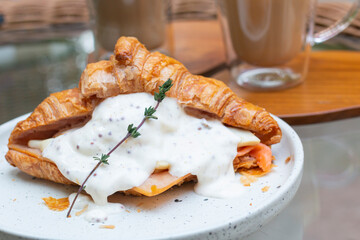 Croissant sandwiches and coffee cups on wooden table. Croissant sandwich with poached egg and smoked salmon on white plate. Sandwich with egg and homemade sauce, cheese and smoked salmon.