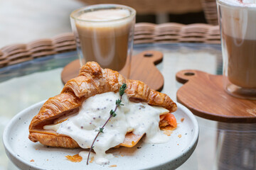 Croissant sandwiches and coffee cups on wooden table. Croissant sandwich with poached egg and smoked salmon on white plate. Sandwich with egg and homemade sauce, cheese and smoked salmon.