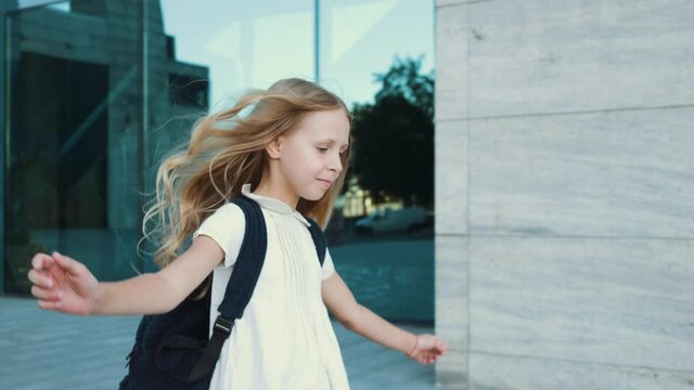 Lonely active little girl with golden hair wears dress and backpack, walks along city street, runs jumps joyfully against background of glass building, rushes elementary school for first grade lesson