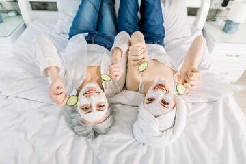 Top view portrait showing two women, senior pretty grandmother and young smiling granddaughter, lying on the bed at home, with facial masks on face, holding cucumber slices. Family spa