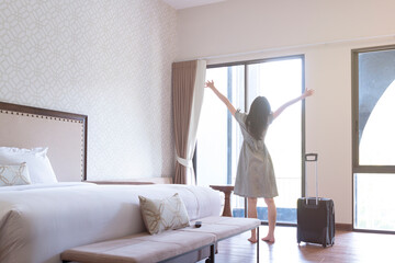 Happy tourist female raised her hands and standing nearly window, looking to beautiful view with her luggage in hotel bedroom. Young woman with suitcase in a hotel room. Happy girl in hotel room.