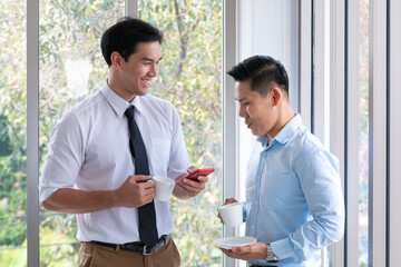 Cheerful business men talking during Coffee break. Businessmen discussing at meeting at office. Business executive man and colleague standing in modern office window discussing after conference.