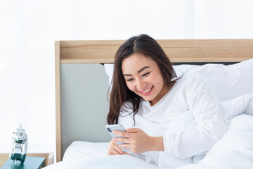 Happy Smiling girl listening music on her smartphone lying on bed. Smiling Asian Girl holding mobile phone on her bed. Using a mobile phone lying on the bed at home. Girl relaxing playing phone in bed