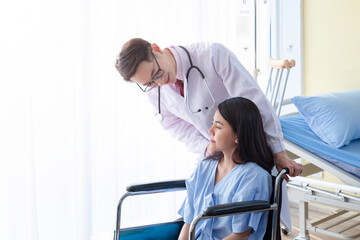 Obraz premium Positive doctor and patient on wheelchair. Female patient being pushed in Wheelchair by Young doctor. Doctor talking to young female patient on wheelchair visiting doctor for regular check.