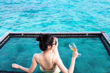 Beautiful young woman with a glass of wine sitting on over-water hammock. Young girl enjoying glass of wine by the sea. Attractive woman enjoying glass of wine. Honeymoon high end travel holiday.