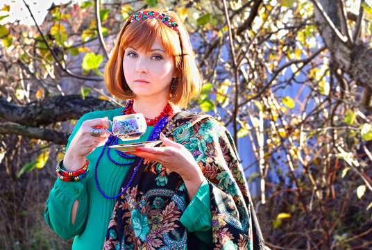 Beautiful young girl with red hair and green eyes in bright clothes is standing with a cup in her hands.