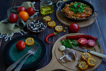 Dark wooden table with a cutting board and a knife, tomatoes, garlic, herbs, corn, a pan with stewed vegetables, beans, hot pepper and serving green plates.