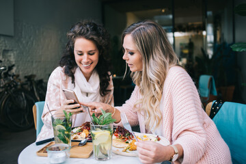 Young blonde and brunette female friends choosing item in web store online using mobile phone during lunch break, pretty caucasian girls looking at common photo on smartphone sitting in cafe