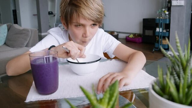 Cute sleepy young boy eating breakfast in kitchen with milk in cereal bowl. Kid eating self-made breakfast with healthy smoothie at home.