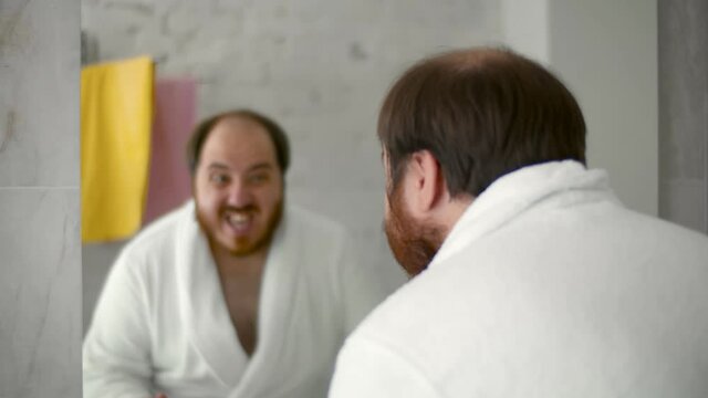 Funny overweight man in bathrobe looking in bathroom mirror and dancing