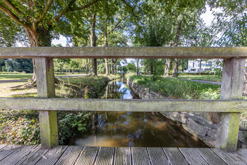 Keutelbeek stream with lush green trees on the shore and the reflection of sunlight in the water seen from the small wooden petonal bridge, sunny summer day in Sittard in South Limburg, Netherlands