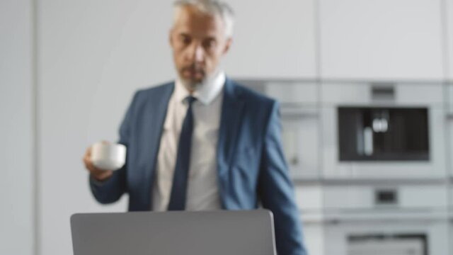 Senior businessman taking coffee from automated coffee machine and working on laptop at home
