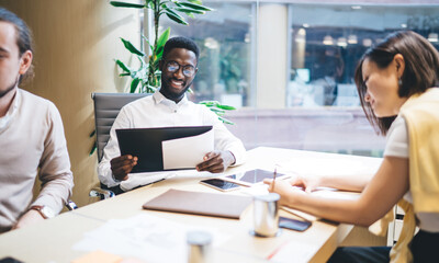 Fototapeta premium Smiling dark skinned male and female diverse group sitting together on meeting table working on strategy and planning creation, prosperous crew of employees satisfied with cooperation on brainstorming
